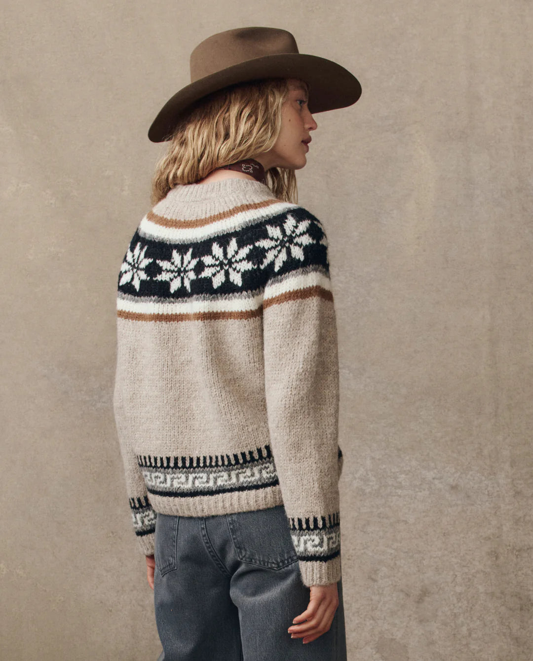 A person in a wide-brimmed brown hat and The Snowflake Fairisle Pullover by The Great Inc.—a beige knit with black and white Nordic motifs—stands sideways against a plain, neutral background.