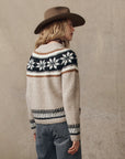 A person in a wide-brimmed brown hat and The Snowflake Fairisle Pullover by The Great Inc.—a beige knit with black and white Nordic motifs—stands sideways against a plain, neutral background.