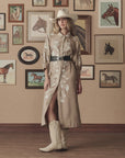 A woman in a cowboy hat, tan embroidered dress, The Great Inc. Tooled Waist Belt, and cream cowboy boots stands on a wooden floor before a wall of framed horse art.