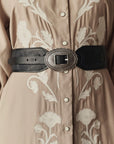 A close-up of a person wearing a beige shirt with white floral embroidery, accessorized with The Great Inc. Tooled Waist Belt featuring an ornate, oval silver buckle.