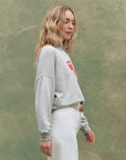 A woman with long blonde hair stands in profile against a textured green background, wearing The Great Inc. Smooth Sailin Sweatshirt and high-waisted white pants with a tan belt.