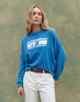 A woman with long blonde hair wears The Great Inc. Smooth Sailin Sweatshirt, a blue vintage top with a seascape graphic, paired with white pants against a textured green background.