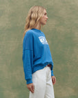 A woman with wavy blonde hair stands in profile against a green background, wearing The Great Inc. Smooth Sailin Sweatshirt with white lettering and white pants.
