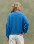 A person with wavy blonde hair models The Great Inc.'s Smooth Sailin Sweatshirt in bright blue and white pants, standing with their back to the camera against a muted green background.