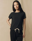 A woman with long dark hair stands against a beige background, wearing The Great Inc.’s The Little Tee in black and black pants with a white drawstring, gazing at the camera with a relaxed expression.