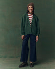 A person stands smiling against a green background, wearing The Brushed Melange Cardigan by The Great Inc. over a red-and-white striped shirt, dark wide-leg jeans, brown shoes, a patterned scarf, and a knitted brown beanie.