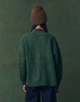 A person faces away wearing The Great Inc. Brushed Melange Cardigan in chunky green, paired with dark blue pants and a brown knit beanie, set against a dark green background.