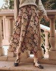 A person in a green "Broncos" sweatshirt and The Great Inc.'s "The Canary Skirt," which features a floral pattern and smocked waist, stands by a stone balustrade. Their face is hidden, but they wear lace-trimmed socks and black-and-white oxford shoes. Trees form the serene backdrop.