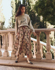A person stands outdoors on a stone pathway with balustrades and trees, wearing a gray sweatshirt with text, The Great Inc.'s The Canary Skirt, and black-and-white shoes. The charming setting resembles a garden or park.