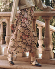 A person stands on a stone terrace, wearing The Great Inc.'s Canary Skirt—a floral design with a smocked waist—paired with a cream sweater featuring writing and vintage-style shoes. The backdrop showcases lush greenery and ornate stone railings.