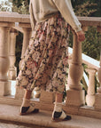 A person in The Great Inc.'s Canary Skirt, cream socks, and brown shoes stands by a stone balustrade. The background is lush with greenery, suggesting an outdoor setting.