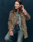 A woman sits against a dark background, smiling as she touches her hair. She wears The Car Coat by The Great Inc. in crushed faux fur leopard print, layered over a denim jacket and red-and-white striped shirt with blue jeans.