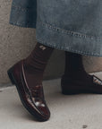 The Great Inc. Monogrammed Trouser Sock in brown, styled with loafers and a denim skirt on concrete.