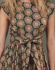 A person with blonde hair wears The Tulsa Dress by The Great Inc., showcasing red and green circular prints on a dark base, complemented by cap sleeves, trapunto stitching, and a waist-tied bow.