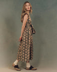 A woman stands against a muted backdrop wearing The Great Inc.'s The Tulsa Dress, featuring a vintage print and an attached belt. Her long hair flows as she pairs socks with two-tone shoes, gazing slightly upwards thoughtfully.