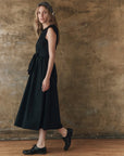 A woman stands sideways against a textured brown wall, wearing The Great Inc.’s Tulsa Dress—a sleeveless black midi with a waist seam—paired with black oxford shoes and a vintage bandana as a headscarf.