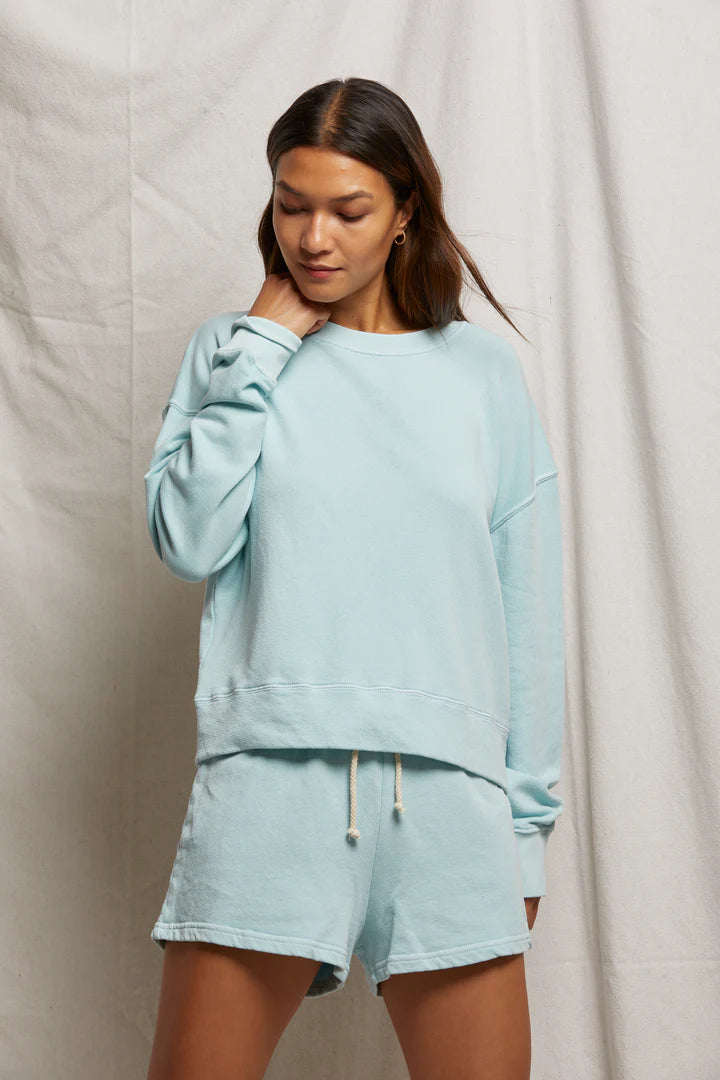 A woman wearing the Perfectwhitetee "Layla Terry Short" light blue sweatshirt and lounge shorts set, made of soft French terry cotton, stands against a beige fabric backdrop, gently touching her neck and looking down with a gentle expression.