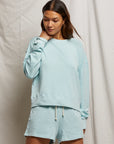 A woman wearing the Perfectwhitetee "Layla Terry Short" light blue sweatshirt and lounge shorts set, made of soft French terry cotton, stands against a beige fabric backdrop, gently touching her neck and looking down with a gentle expression.