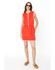 A person in The Terry Dress by Kule, featuring a sleeveless design with white trim and front zipper closure, stands against a white background wearing sunglasses and brown sandals.