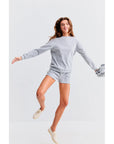 A person jumps joyfully in a light gray sweatshirt and Kule's Terry Venus shorts with an elastic waist, holding a gray jacket. They wear light-colored shoes, all against a plain white background.