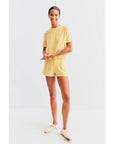A person stands smiling in a minimalist, light-filled space, wearing Kule's matching cotton-blend yellow T-shirt and The Terry Venus short with an elastic waist, paired with cream-colored sneakers.