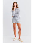 A long-haired person smiles with one hand on their chin, wearing Kule's The Terry Franny sweatshirt in light grey and matching shorts, paired with light-colored shoes against a plain white background.