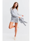 A person stands joyfully on one leg against a light background, wearing Kule's The Terry Franny sweatshirt and shorts. They hold a matching top in one hand, smiling in light sneakers. The outfit's casual charm is enhanced by the raglan sleeves of the gray cotton-blend sweater.