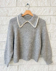 The Great Inc.'s Crochet Collar Pullover is a gray knit, long-sleeve sweater with a scalloped white collar, displayed on a wooden hanger against a white brick wall.