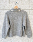 The Great Inc.'s The Crochet Collar Pullover is a gray, relaxed-fit knitted sweater with long sleeves, a high wide collar, and a decorative white crochet trim, shown hanging on a wooden hanger against a white brick wall.