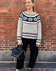 A woman smiles softly to the side against a brick wall, wearing The Great Inc.'s Snowflake Fairisle Pullover in beige with black and white patterns, styled true to size with black pants, shoes, and a handbag.