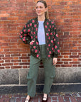 A person stands before a brick wall, wearing The Great Inc.'s "The Zip Front Plush Bomber" in army green with a rose pattern, styled with a white tee, green pants, burgundy flats; hair tied back and one hand on their hip.
