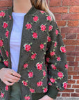 A person stands against a brick wall wearing The Great Inc.'s The Zip Front Plush Bomber in army green with a red and white rose pattern, paired with a white T-shirt and green pants, hand near the zipper.
