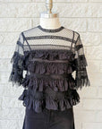 The Winter Mesh Flounce Top by The Great Inc. is a black sheer lace blouse with a sheer upper bodice, horizontal ruffled layers, and intricate woven lace details, displayed on a mannequin against a white brick wall.