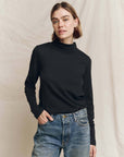 The Great Inc. Turtleneck in beige, styled with blue jeans against a neutral fabric backdrop, hand casually in pocket.