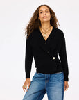 A woman with long curly hair wears the Jackie Cardigan by Kerri Rosenthal—a black V-neck cardigan with buttons—paired with blue jeans and layered gold necklaces, standing confidently against a plain white background.