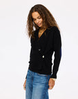 A woman with long curly hair, wearing the Kerri Rosenthal Jackie Cardigan in black and blue jeans, stands against a plain light background, looking down with a relaxed expression.