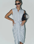 A woman wears THE SHIRT’s Sleeveless Wrap Front Dress in light blue stripes with a waist tie and No Gape button, sunglasses, and holds a black handbag against a plain white background.