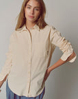 A woman with long light brown hair wears the Super Relaxed Boyfriend shirt by THE SHIRT in beige cotton poplin with rolled sleeves and blue pants, standing against a plain light background, looking relaxed.