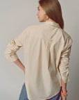 A woman with long brown hair wears THE SHIRT’s Super Relaxed Boyfriend in cream cotton poplin and blue pants, facing away from the camera. She accessorizes with gold hoop earrings and colorful bracelets on her right wrist.