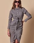 A woman wearing THE SHIRT's The Wrap Front Dress—a black-and-white checkered, long sleeve shirt dress with a waist tie—stands against a beige background.