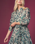 A woman poses against a maroon background, wearing THE SHIRT's Jenica Dress with No Gape button technology, featuring a green and black leopard print and a cinched, flared skirt.