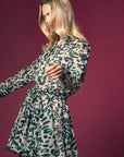 A woman with long blonde hair models the Jenica Dress by THE SHIRT, a patterned shirtdress with green and black spots and a braided rope belt, standing against a solid maroon background.