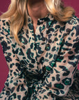A woman with wavy blonde hair models THE SHIRT's Jenica Dress, a green, black, and beige leopard print shirtdress featuring a braided rope belt and No Gape button technology, seated against a deep pink background with her face partially out of frame.