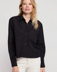 A woman with long blonde hair wears THE SHIRT's The LS Boxy Crop, a black button-up, and white pants, standing against a plain light gray background.