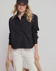 A woman in THE SHIRT's The LS Boxy Crop—a black button-up—white pants, and a black baseball cap stands against a plain light background, holding a tan sweater in both hands.