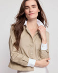 A woman with long brown hair wears The LS Boxy Crop by THE SHIRT, a beige button-up with white cuffs and collar featuring No Gape technology, standing against a plain light background and smiling softly.