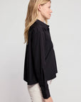 A woman with long blonde hair stands in profile wearing THE SHIRT's The LS Boxy Crop—a loose-fitting black button-up blouse—paired with light-colored pants against a plain light background.