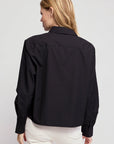 A woman with blonde hair is seen from the back, wearing THE SHIRT’s The LS Boxy Crop—a black, long-sleeve button-up—paired with off-white pants against a plain light grey background.