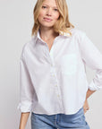 A woman with blonde hair wears THE SHIRT’s The LS Boxy Crop, a classic white cropped shirt with rolled-up sleeves, paired with blue jeans. She stands against a light background and smiles softly at the camera.