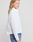A woman with blonde hair wears THE SHIRT's LS Boxy Crop, a white women’s button-up, paired with blue jeans, standing in profile against a plain light background.
